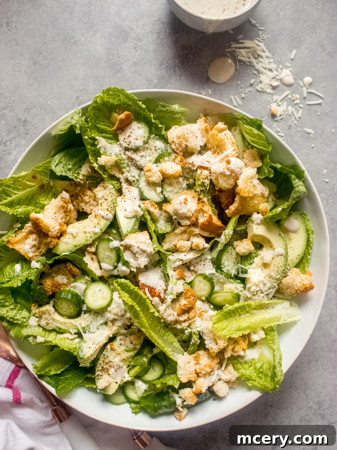 Lush Avocado Caesar 2 A vibrant Caesar salad with generous slices of avocado and fresh cucumber, elegantly presented in a white bowl, ready to be enjoyed.
