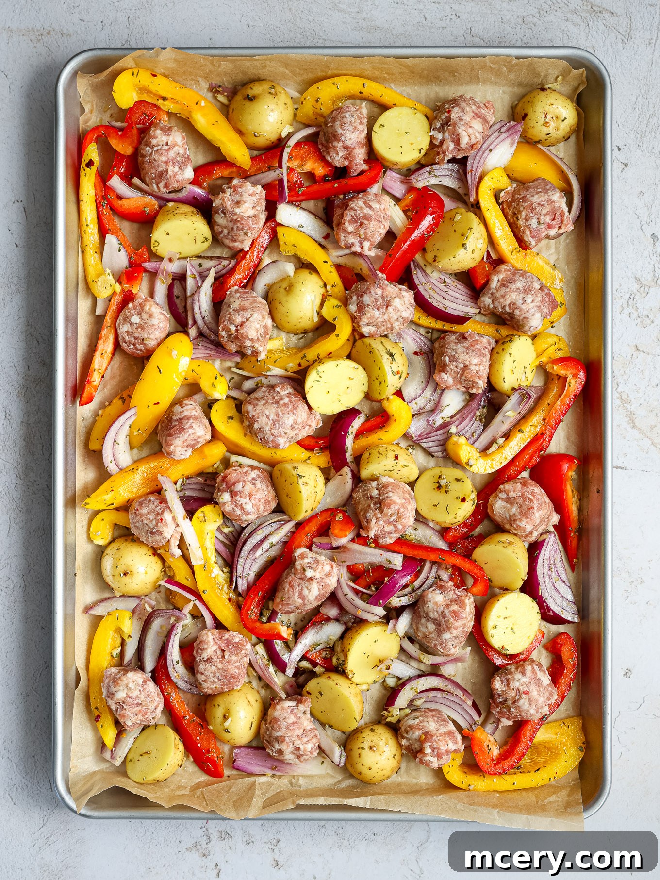 Halfed baby gold potatoes and raw Italian sausage chunks spread evenly on a parchment-lined baking sheet, ready for roasting.