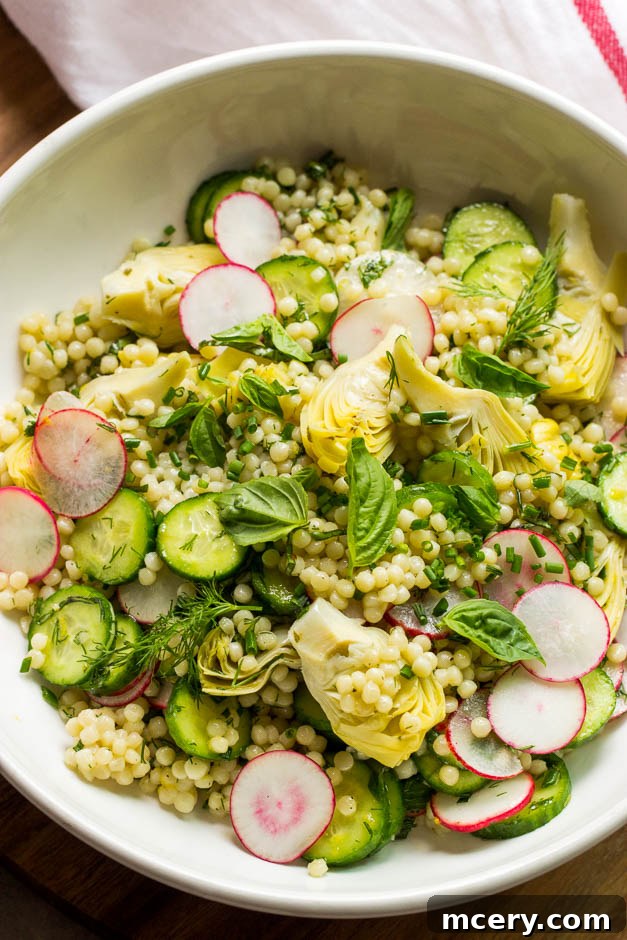 Jeweled Israeli Couscous Salad 2 Close up Israeli couscous salad with marinated artichokes in white bowl, garnished with fresh herbs.