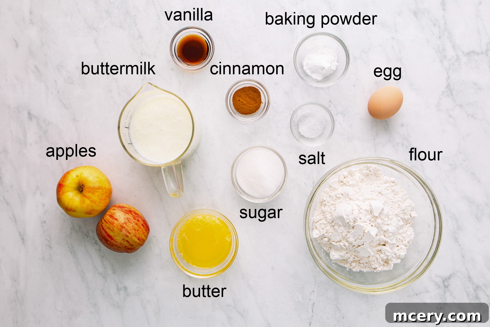 A flat lay of fresh ingredients for apple cinnamon pancakes, including apples, flour, eggs, butter, buttermilk, sugar, baking powder, cinnamon, and vanilla extract.