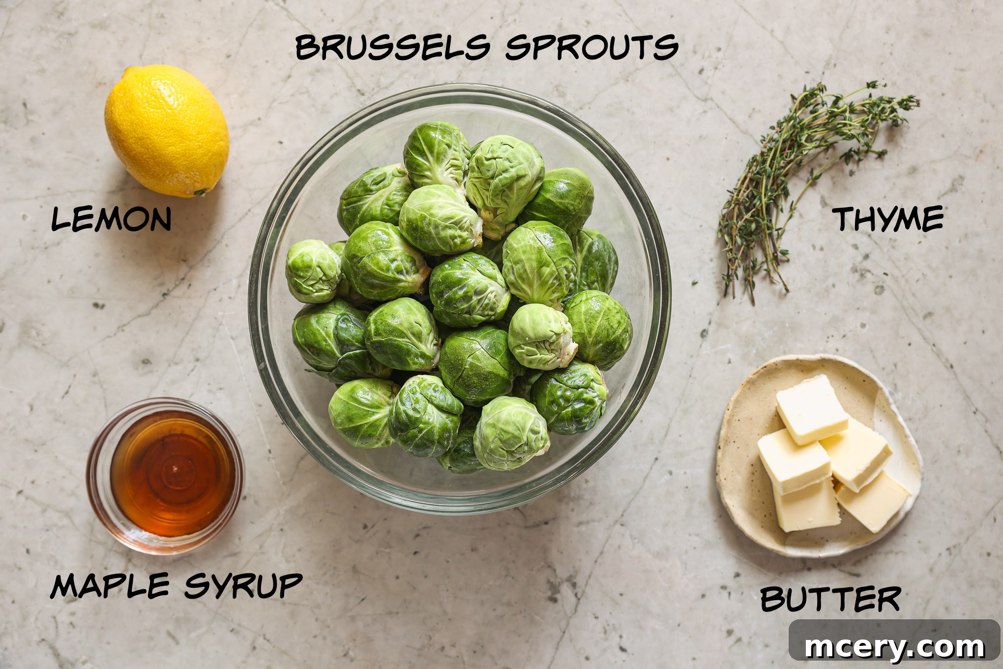 A visual spread of fresh Brussels sprouts, a bottle of maple syrup, a stick of butter, fresh thyme sprigs, and a lemon, laid out as ingredients.