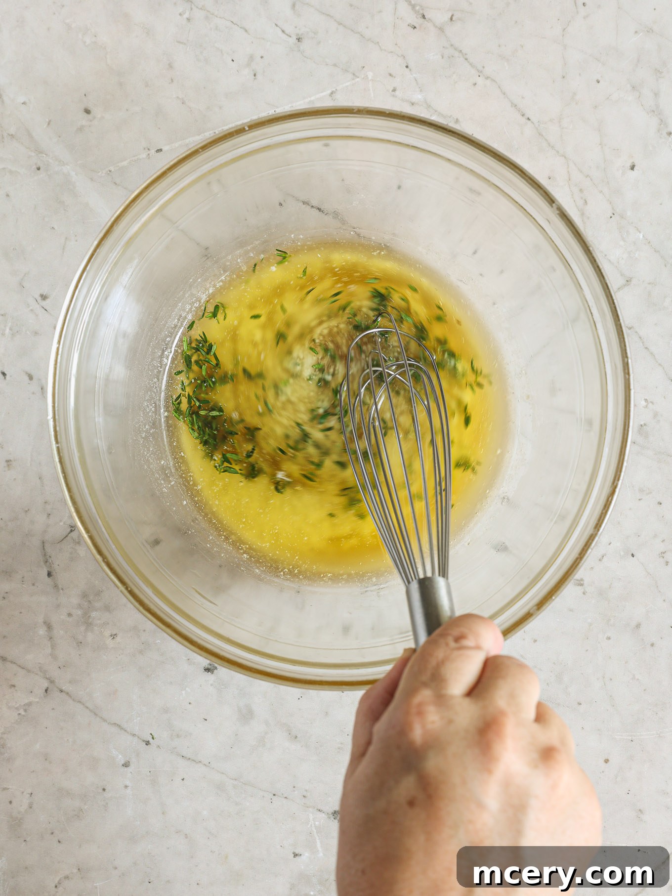 A glass bowl containing a rich mixture of melted butter, golden maple syrup, and fresh green thyme sprigs, ready for whisking.