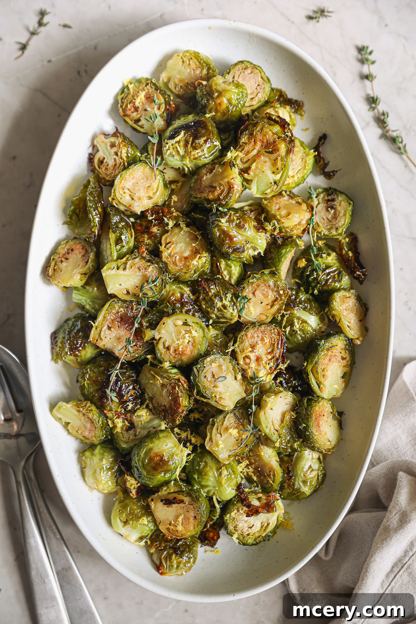 Close-up of maple roasted Brussels sprouts garnished with bright yellow lemon zest, served on a textured white serving dish.