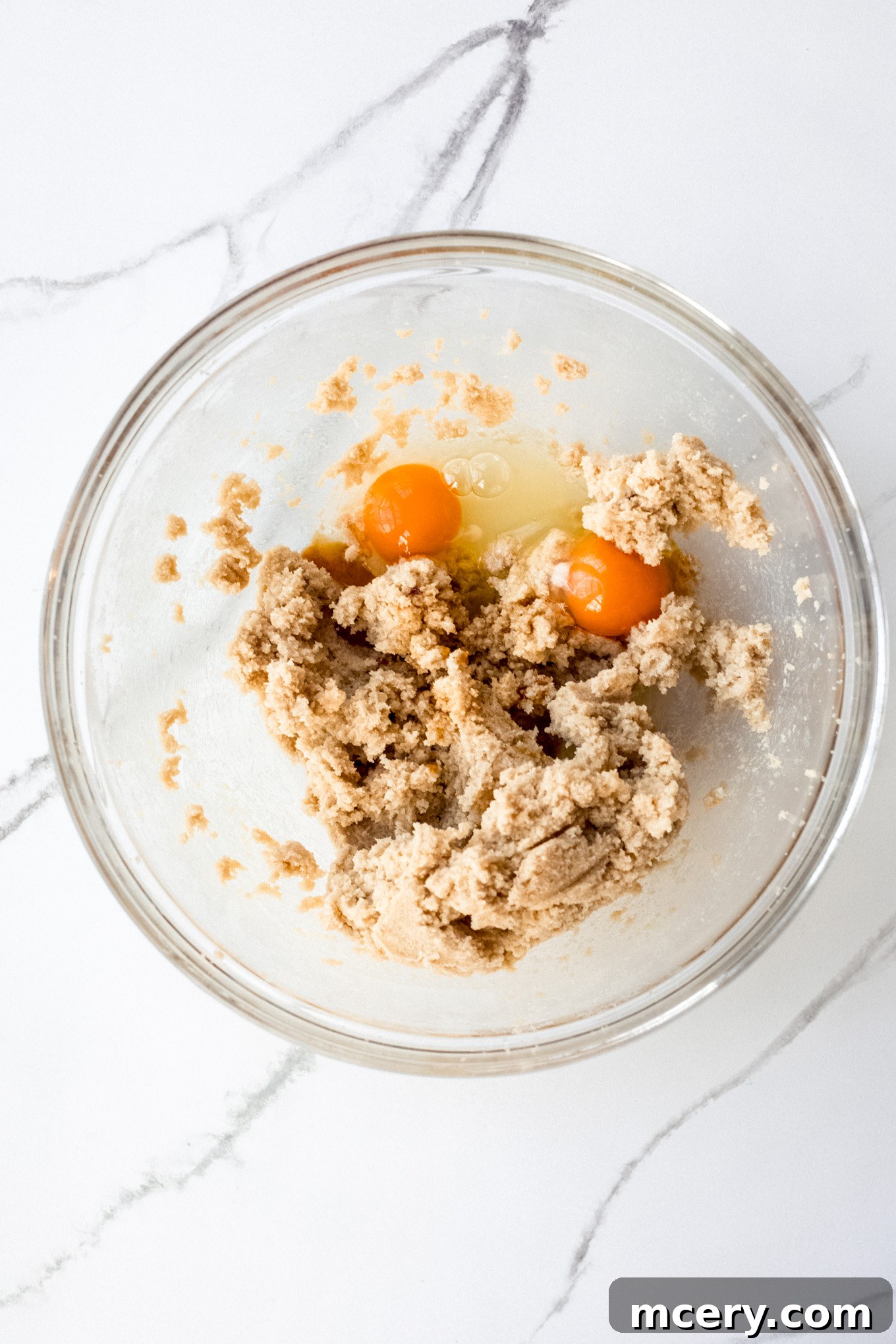 Close-up of butter, brown sugar, cane sugar, and eggs being creamed together in a glass bowl with an electric mixer.