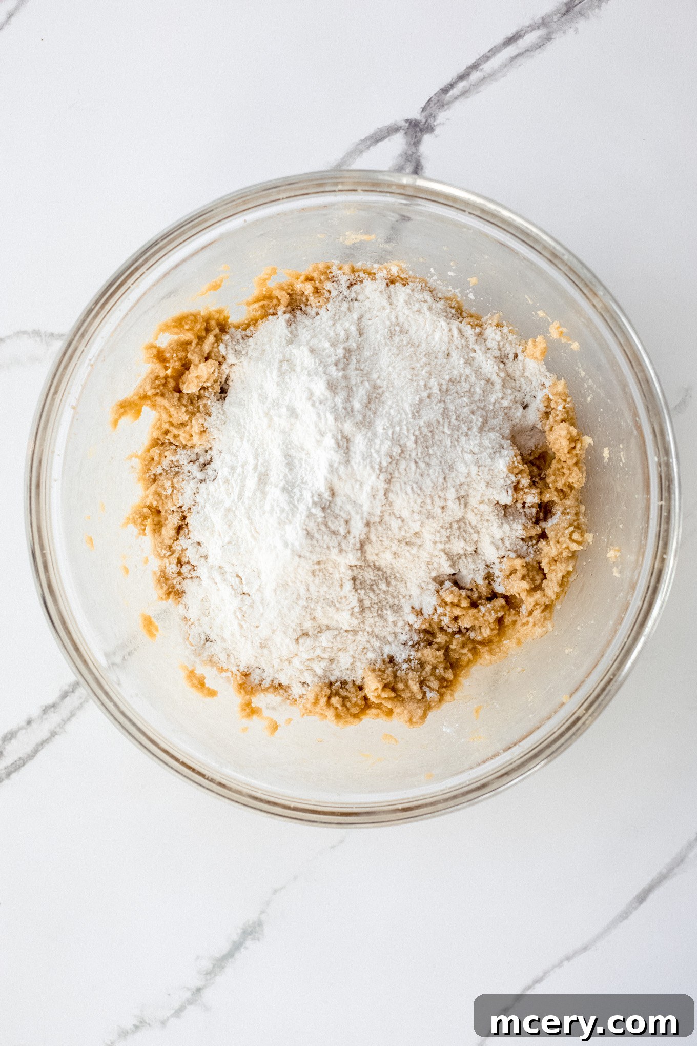 Dry ingredients, including flour, baking soda, and salt, being whisked together in a separate glass bowl before being added to wet ingredients.