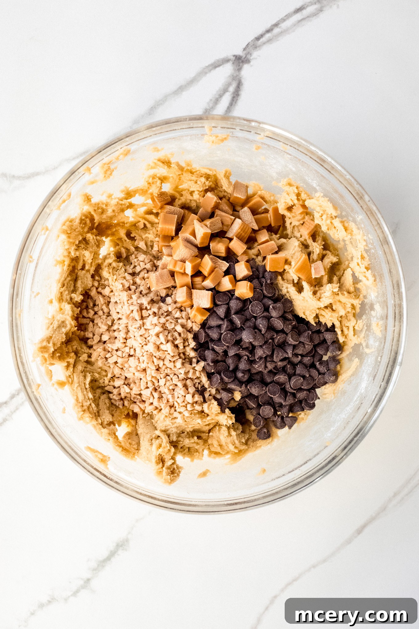 Chopped chocolate chips, toffee bits, and soft caramels resting on top of cookie dough in a glass bowl, ready to be folded in.