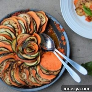 Hearty Sweet Potato Ratatouille 2 A round dish of colorful Sweet Potato Ratatouille, with thinly sliced layers of zucchini, eggplant, and tomato arranged in a spiral pattern. A serving spoon rests on top. A plate with a portion of ratatouille is beside the dish on a gray surface.