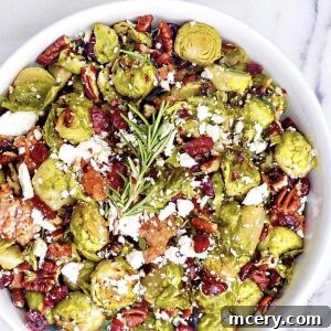 A round white bowl filled with roasted Brussels sprouts and sausage, garnished with pecans, cranberries, and crumbled feta cheese, with a sprig of fresh rosemary.