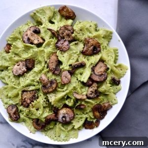 A plate of bow-tie pasta with creamy Vegan Basil Cream Sauce, topped with sautéed mushrooms, served on a white dish. A grey cloth is partially visible on the side against a marble surface.