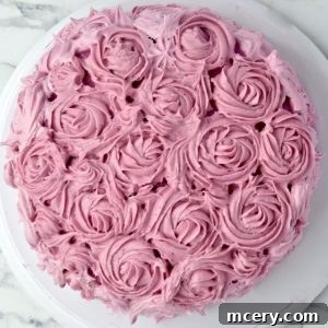 A round cake decorated with intricate pink Pitaya Cream Cheese Frosting roses, placed on a white cake stand. The frosting is textured to resemble blooming flowers, creating an elegant and delicate appearance.