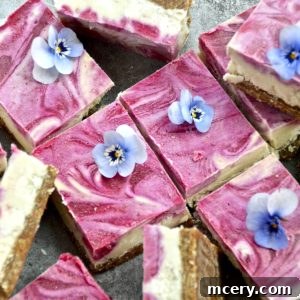 Sliced square pieces of a pink and white swirled Vegan No-Bake Lemon Raspberry Bars topped with edible blue flowers on a gray background. The dessert has a crumbly brown crust.