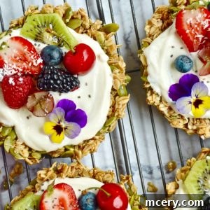 Delicious Granola Tarts topped with creamy yogurt, fresh berries, kiwi slices, chia seeds, and edible flowers, placed on a cooling rack over a marble surface.