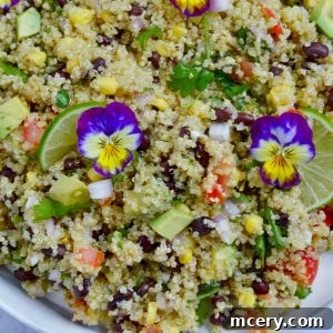 A vibrant Mexican Quinoa Salad with black beans, corn, avocado, tomatoes, and red onion, garnished with lime slices and edible purple flowers. Served in a white bowl on a light marble surface.