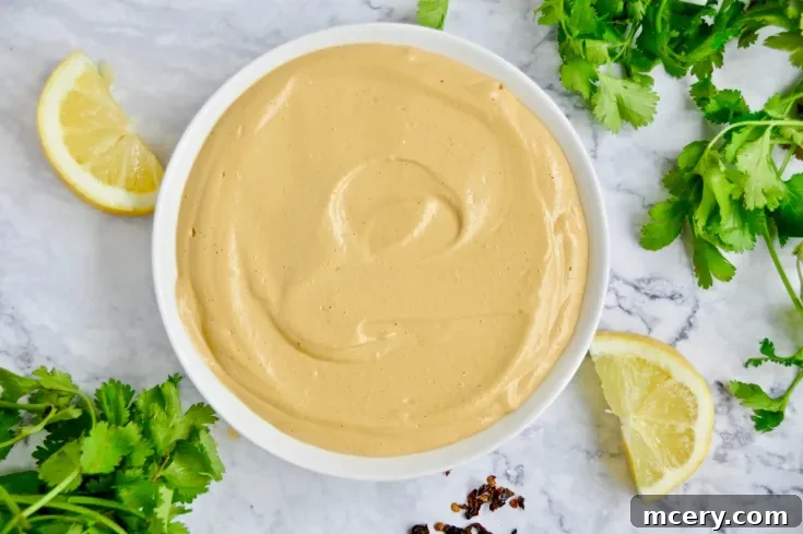 A top-down view of a small bowl filled with smooth, pale orange Chipotle Cashew Cream, surrounded by fresh cilantro leaves and a slice of lime.