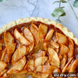 A freshly baked apple pie with a golden crust, topped with glazed apple slices, sits on a marble surface. Green leaves are visible beside the pie, adding a touch of nature to the scene.