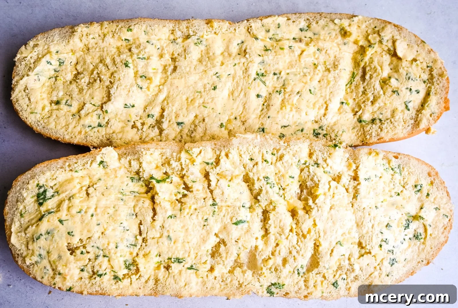 Close-up of a rustic wooden board holding freshly baked cheesy garlic bread. The bread is sliced diagonally, revealing a golden-brown crust and a generous topping of melted cheese and green herbs. A few loose crumbs are visible, suggesting its delightful texture. The background is softly blurred, focusing attention on the appetizing bread.