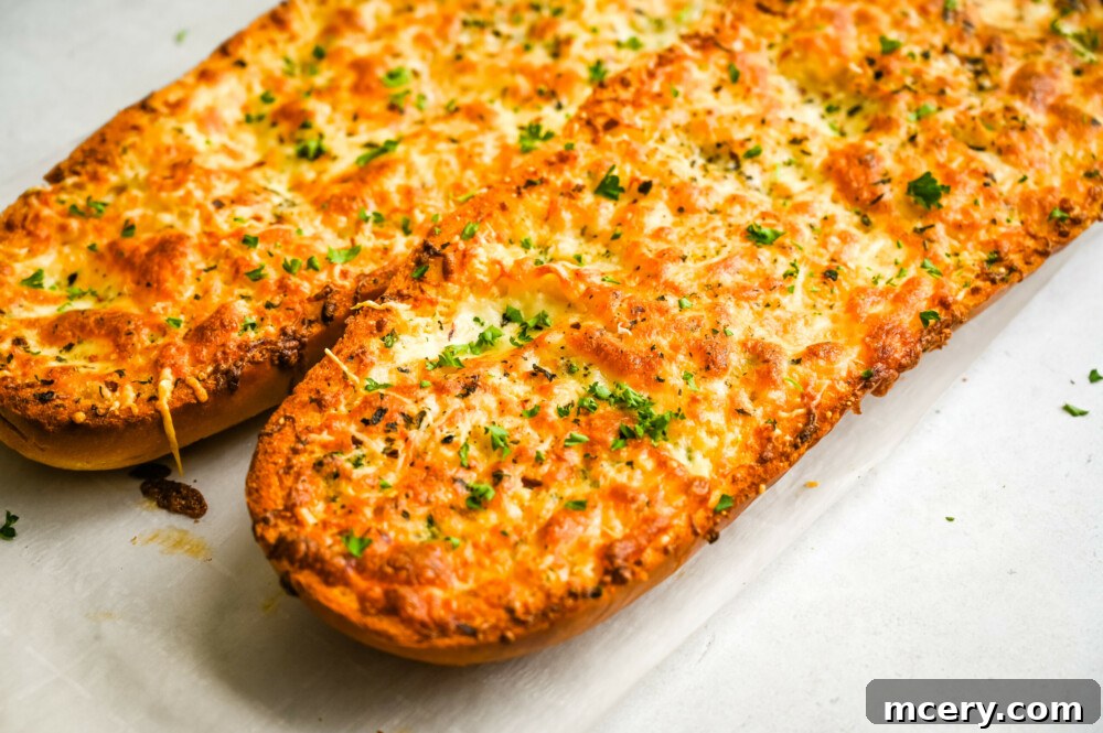 A golden-brown loaf of cheesy garlic bread, freshly baked and glistening with melted cheese and butter. The top is slightly crusty, hinting at its crunchy texture. It rests on a textured white background, possibly parchment paper, which emphasizes its warmth and homemade appeal.