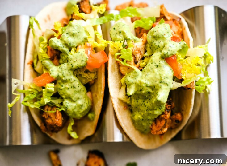 Close-up of Chicken Tacos with Creamy Cilantro Sauce in a metal holder. The taco is filled with grilled chicken, chopped lettuce, diced tomatoes, and topped with a generous serving of creamy cilantro sauce. The bright colors and fresh ingredients make it look appetizing.