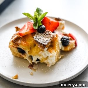 A slice of Berries and Cream French Toast Bake topped with fresh strawberries, blueberries, a sprig of mint, and a dusting of powdered sugar sits on a white plate.