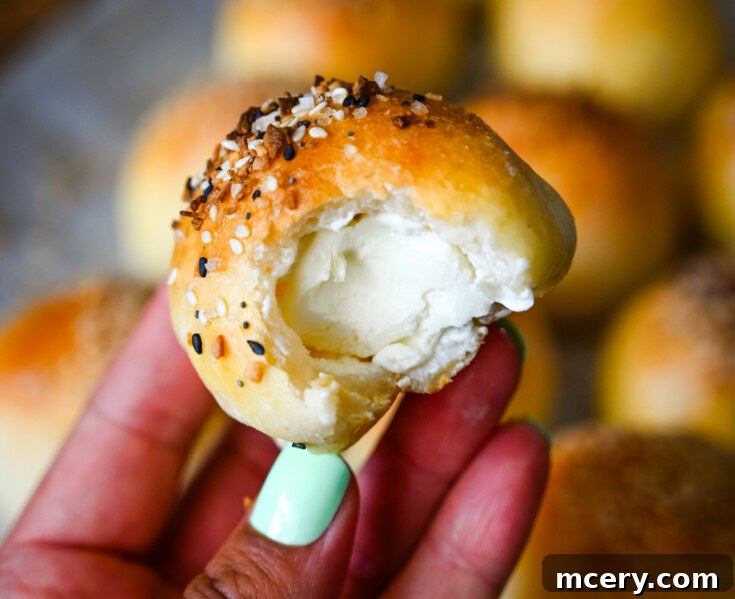 A hand with mint green nail polish holds a bite-sized stuffed bagel bite with a fluffy white cream cheese filling. The bagel bite is topped with sesame and other seeds. Several more similar rolls are blurred in the background.