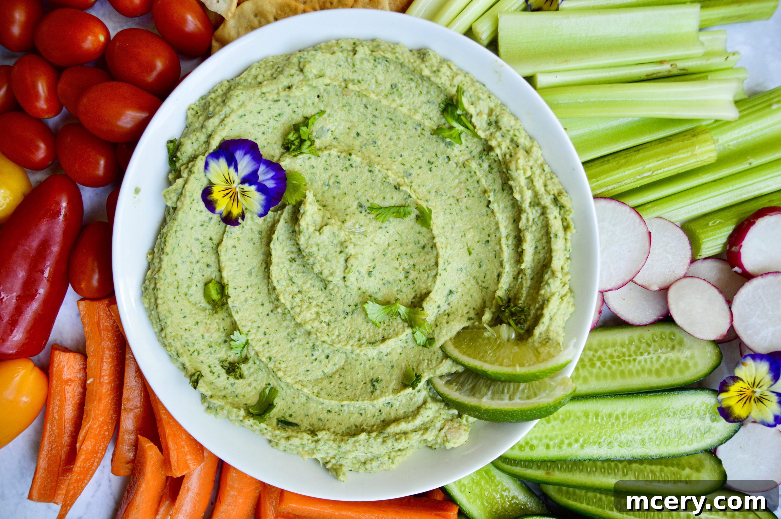 Zesty Cilantro Jalapeño Hummus 3 A close-up of creamy cilantro jalapeño hummus in a bowl, garnished with a sprig of fresh cilantro, sitting on a rustic wooden board next to a colorful array of fresh vegetables and crispy crackers for dipping.
