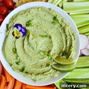 A platter with a bowl of creamy Cilantro Jalapeño Hummus garnished with lime slices and an edible flower, surrounded by sliced cucumber, radish, celery, carrot sticks, cherry tomatoes, crackers, and bell pepper slices on a marble surface.
