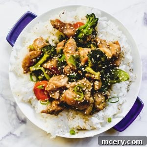 Quick and Flavorful Teriyaki Chicken Rice Bowls 6 A Teriyaki Chicken and Rice Bowl atop white rice. The dish includes broccoli, carrots, and green onions, and is garnished with sesame seeds. The bowl has purple handles and rests on a light marble surface.
