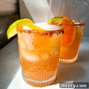 Two glasses of refreshing Grapefruit Margaritas, with a chili powder-rimmed glass, garnished with lime wedges and slices of citrus inside. The drink has an orange hue and is placed on a countertop with sunlight streaming in. Another similar glass is out of focus in the background.