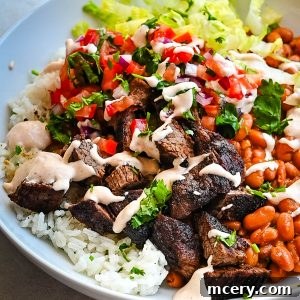 Smoky Chipotle Steak Bowls 6 A white bowl contains a Steak Burrito Bowl on cilantro rice, topped with diced tomatoes, chopped lettuce, cooked beans, and drizzled with creamy sauce. Garnished with fresh cilantro.