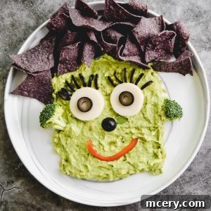 A plate of FrankenFine Guacamole shaped like a Frankenstein with black olive eyes, a red pepper smile, broccoli ears, and tortilla chips as hair, arranged on a marble surface.