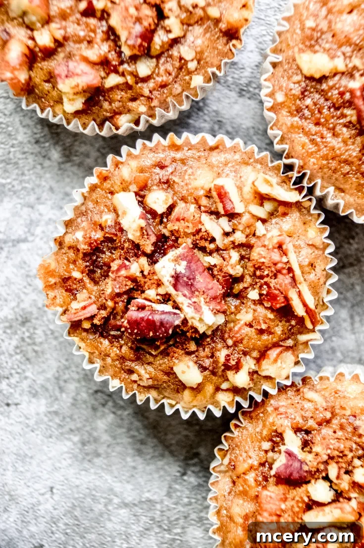 Four Vegan Banana Cinnamon Muffins topped with chopped nuts, arranged on a textured gray surface. The muffins are golden brown with a crumbly texture, and each is in a white paper liner.