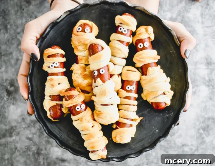 A black plate held by two hands contains five sausages wrapped in pastry to resemble mummies. Each has small white eyes with black pupils, giving a playful and spooky appearance. The background is a textured gray surface.