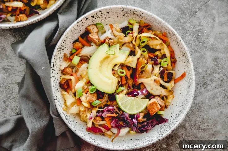 Egg Roll in a Bowl with rice noodles, sliced avocado, lime wedge, scallions, and mixed vegetables, including purple cabbage and carrots, garnished with tofu chunks. A gray napkin partially visible on the side.