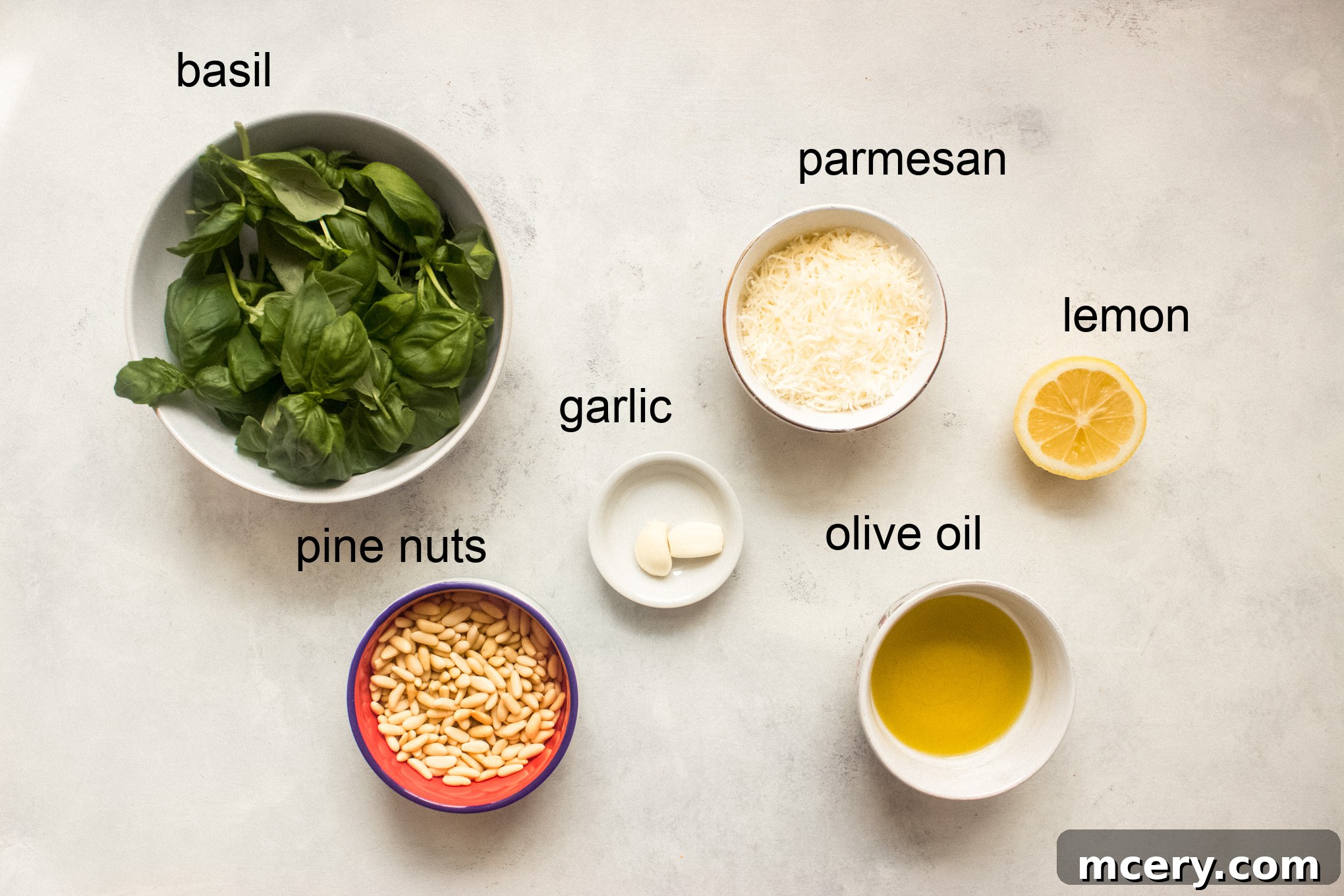 A beautifully arranged spread of fresh ingredients on a light wooden surface, including a generous bunch of green basil leaves, a small bowl of golden pine nuts, a wedge of Parmesan cheese, several garlic cloves, a bottle of premium olive oil, and bright yellow lemons, all set for making homemade basil pesto.