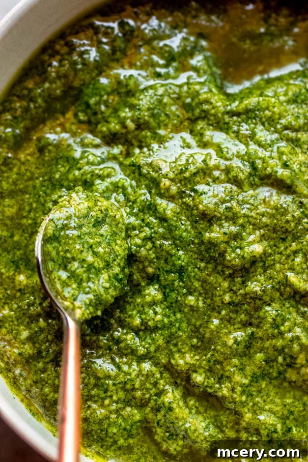 A close-up shot of vibrant green basil pesto in a small glass jar, adorned with fresh basil leaves and a scattering of pine nuts, resting on a textured wooden surface, emphasizing its homemade appeal and natural ingredients.