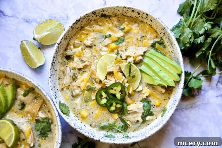 A bowl of creamy white chicken chili is garnished with lime slices, sliced avocado, jalapeño peppers, and fresh cilantro. Another bowl with the same contents is partially visible. Lime wedges and a bunch of fresh cilantro are placed alongside the bowl.