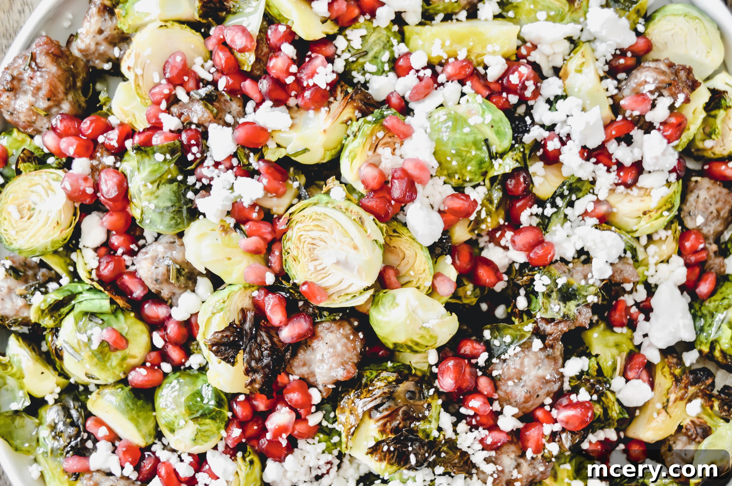 A beautifully presented bowl of roasted Brussels sprouts and Italian sausage, garnished with fresh pomegranate seeds, feta, and pistachios, ready to be served at a festive gathering.