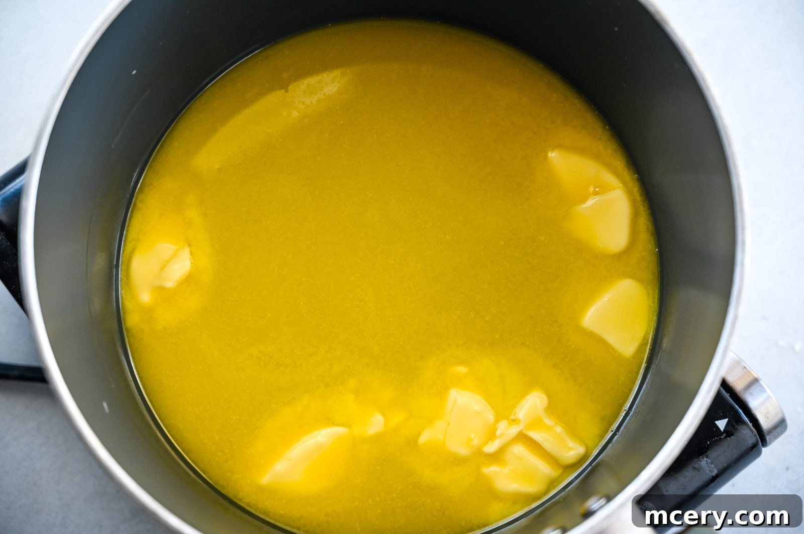 Pratik İç Harcı Tarifi: Şükran Günü Sofralarının Yıldızı 6 A close-up view of a black pot on a stovetop containing melted butter with some solid chunks floating in it. The melted butter is evenly spread across the bottom of the pot, creating a smooth surface with hints of texture from the floating chunks.