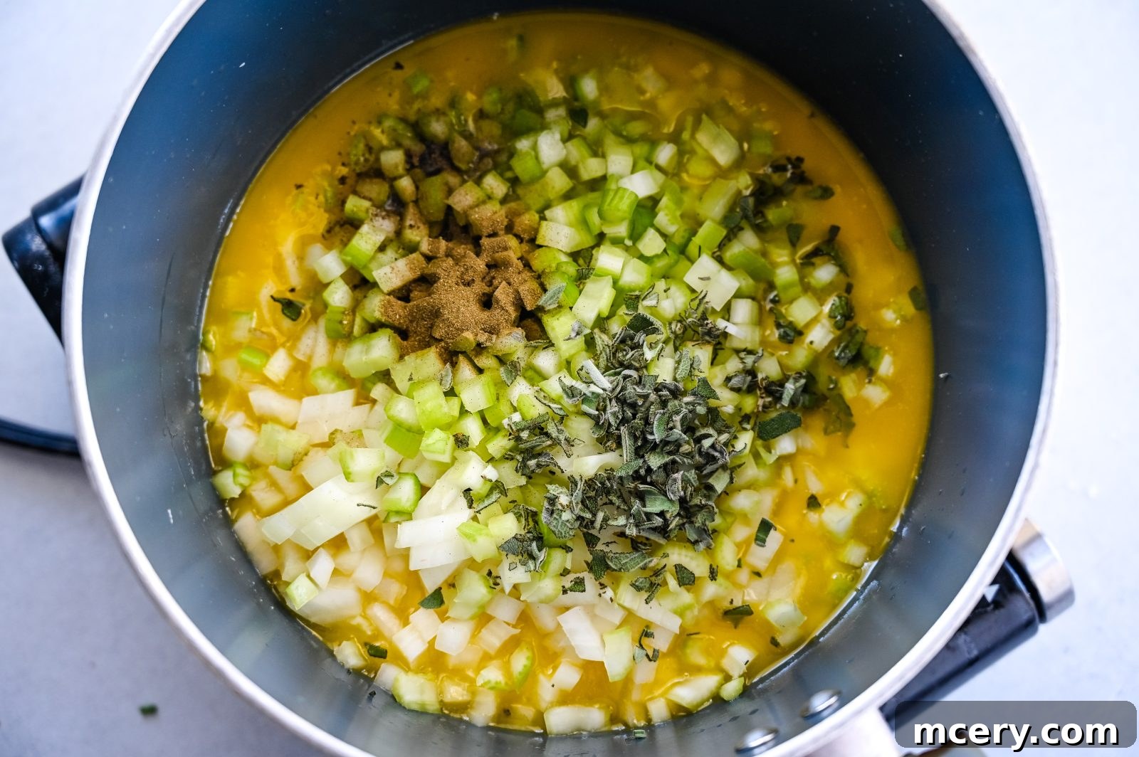 Pratik İç Harcı Tarifi: Şükran Günü Sofralarının Yıldızı 7 A pot on a stove contains a mixture of ingredients including chopped onions, diced celery, butter, and various herbs and spices. The mixture is being prepared for making Easy Stuffing.