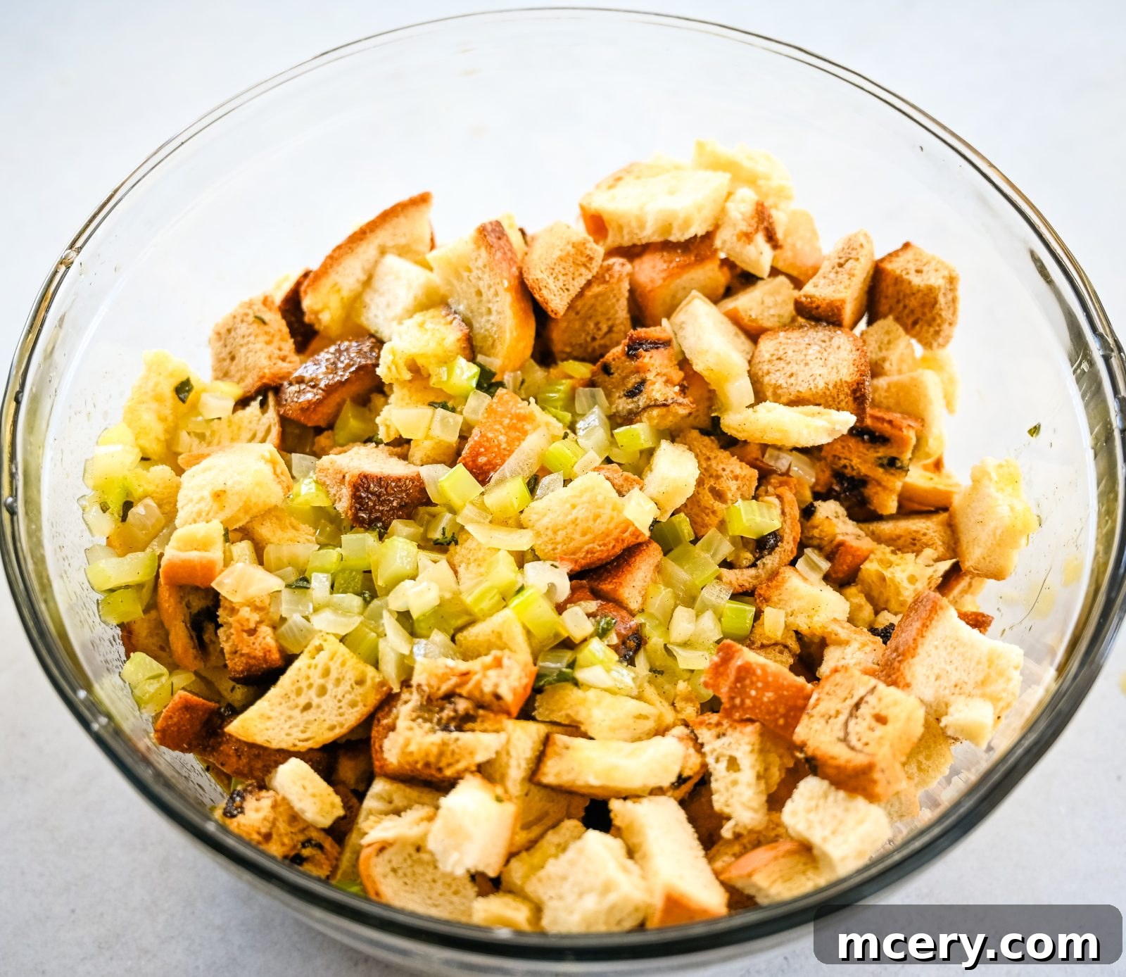 Pratik İç Harcı Tarifi: Şükran Günü Sofralarının Yıldızı 9 A large clear glass bowl filled with cubed bread pieces and diced celery, creating the base for a stuffing mix. The mixture appears fresh and undressed, ready for further preparation and seasoning.