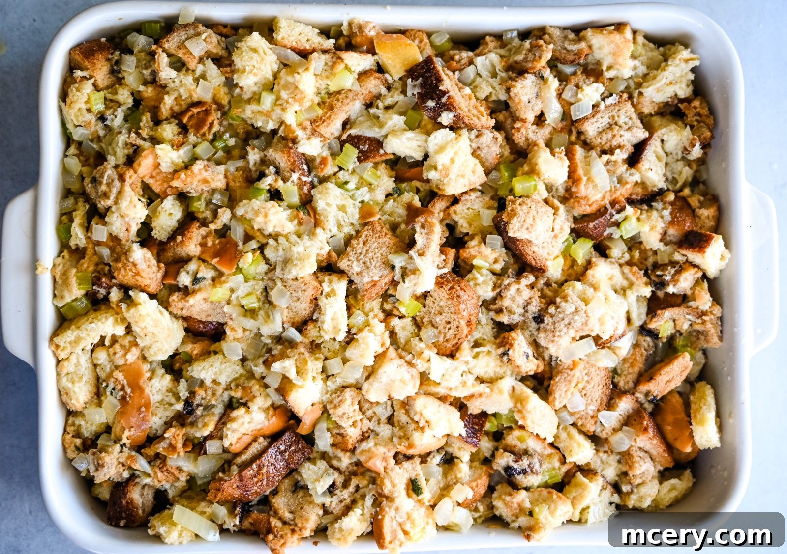 Pratik İç Harcı Tarifi: Şükran Günü Sofralarının Yıldızı 10 A white baking dish filled with unbaked Easy Stuffing made of cubed bread, chopped celery, onions, and herbs. The stuffing appears golden brown and slightly crispy on top, with bits of toasted bread and cooked vegetables clearly visible.