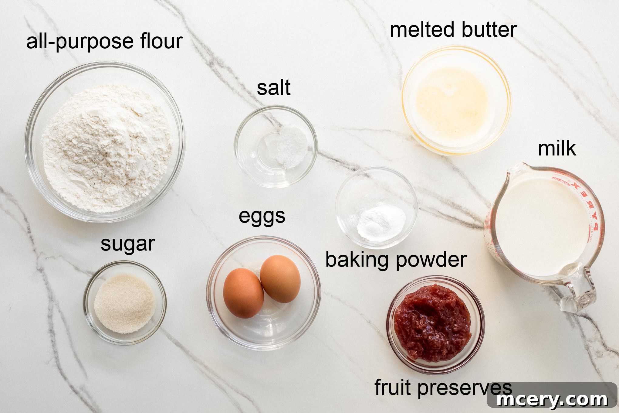 A collection of fresh ingredients for Danish pancakes, including flour, eggs, milk, butter, and fruit preserves, laid out on a kitchen surface.