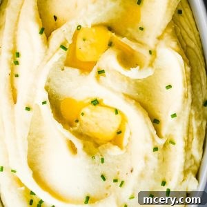 A dish filled with creamy mashed potatoes, topped with melted butter and garnished with finely chopped chives. The mashed potatoes appear smooth and velvety, with shallow dips holding pockets of melted butter, and the chives add a touch of green.