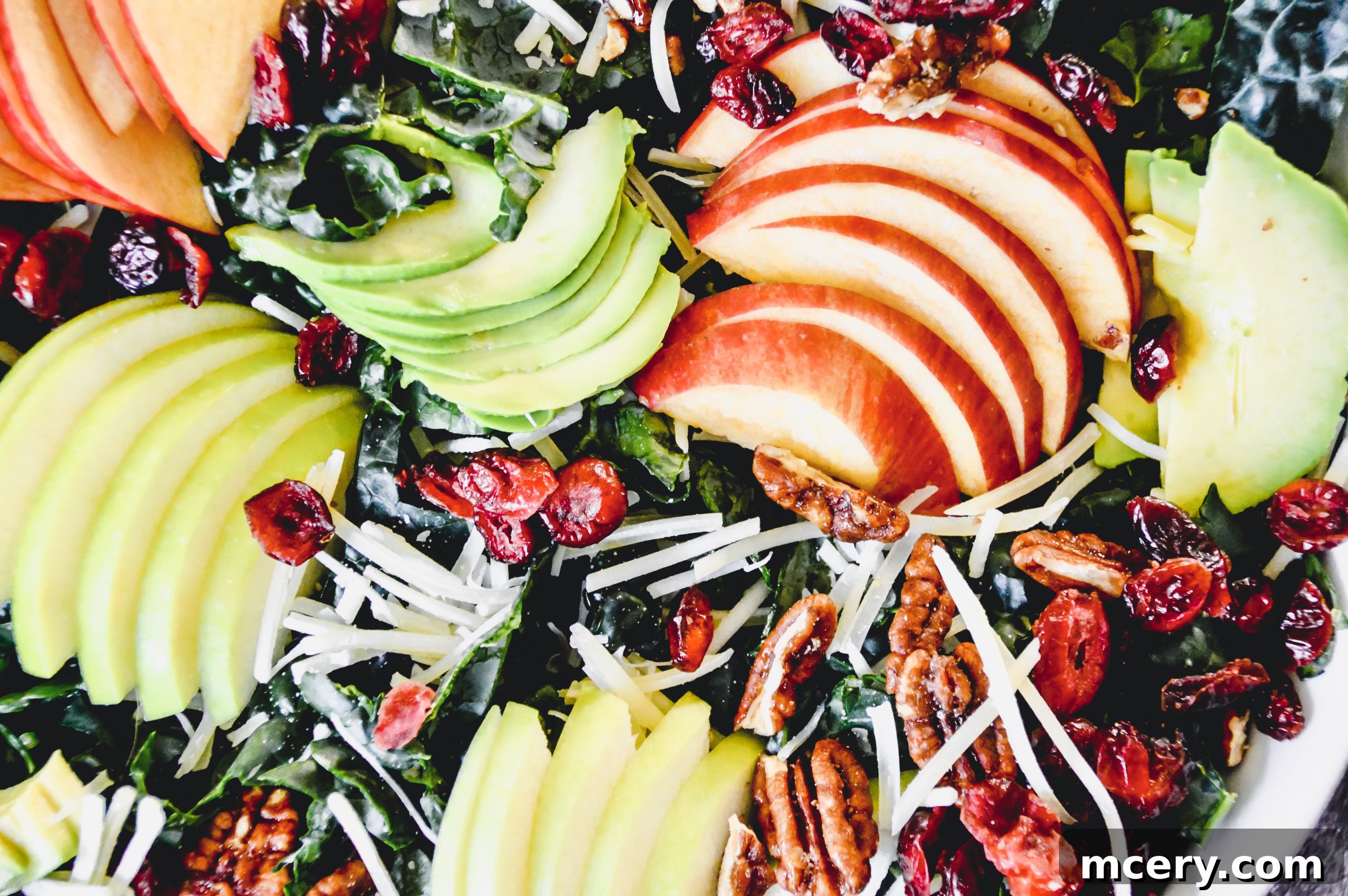 A large bowl of vibrant Tuscan Kale Salad, showcasing fresh green kale, sliced red and green apples, avocado, and sprinkled with various toppings.