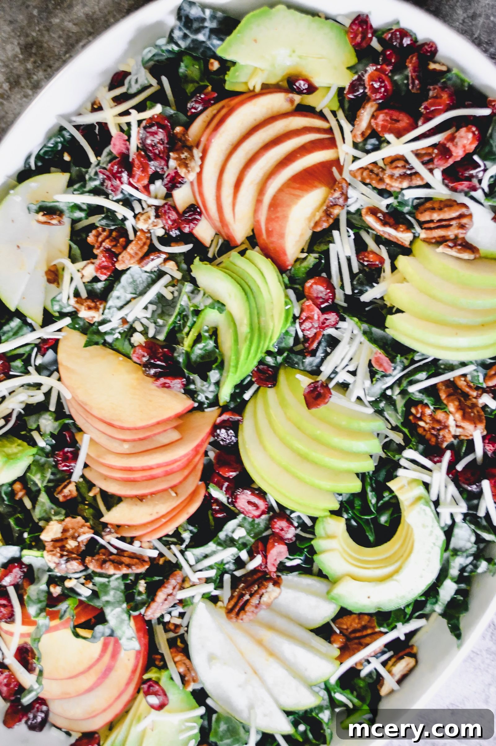 A vibrant Tuscan Kale Salad with Apples in an oval dish with kale, sliced apples and avocados, cranberries, nuts, shredded cheese, and croutons on a textured gray background.