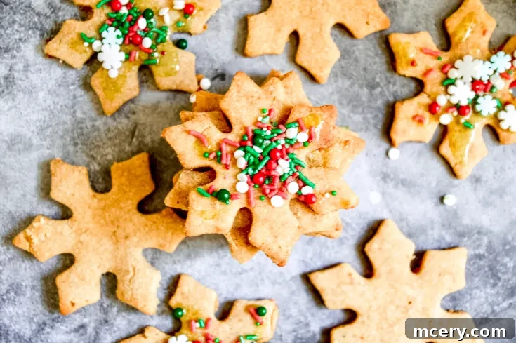 Snowflake-shaped Paleo Cut-Out Skinny Sugar Cookies topped with red, green, and white sprinkles are scattered on a gray surface. One cookie is stacked on top, highlighting its colorful sprinkles.