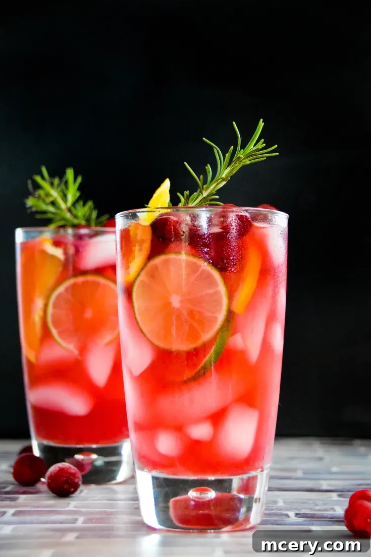 Two glasses of cranberry-infused skinny Jingle Juice Cocktail with ice, garnished with lime, orange slices, cranberries, and rosemary sprigs. Styled on a tiled surface, the vibrant colors and fresh ingredients offer an appealing and refreshing presentation.
