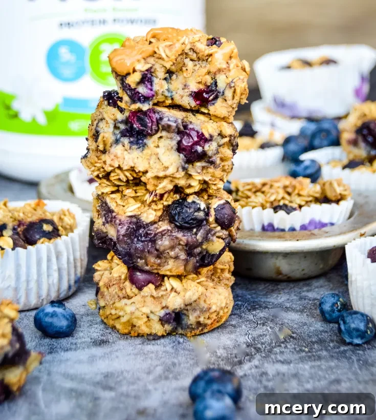 A stack of four Blueberry Protein Oatmeal Cups topped with peanut butter, surrounded by loose blueberries and additional muffins in paper liners. A container of protein powder is blurred in the background.