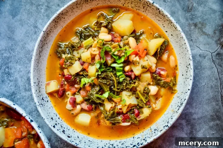 A bowl of vibrant Instant Pot Bean Potato & Kale Vegan Soup with red kidney beans, white beans, diced potatoes, leafy greens, and fresh basil garnish. The broth is a rich, warm orange color, served in a white speckled bowl on a gray surface.