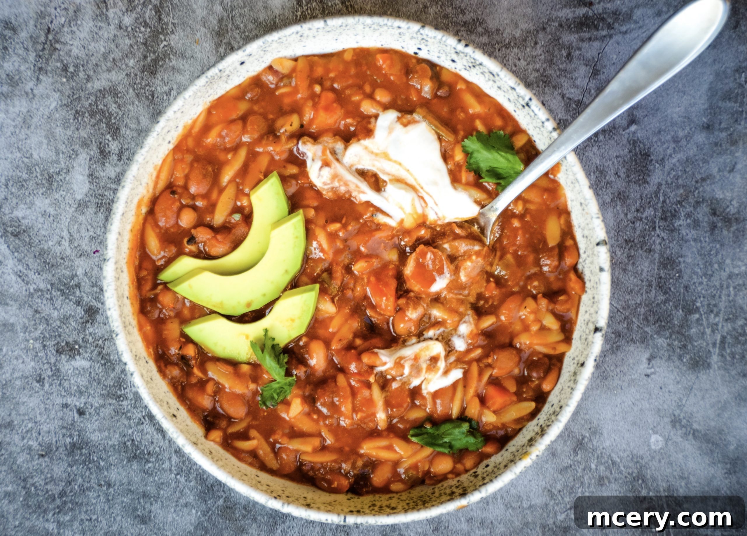 A close-up shot of a steaming bowl of Instant Pot Vegan 10 Bean Taco Soup, ready to be served. The rich, red broth is visible, along with a variety of beans, diced tomatoes, and orzo, promising a comforting and flavorful meal. Garnished with a swirl of vegan sour cream and fresh herbs.
